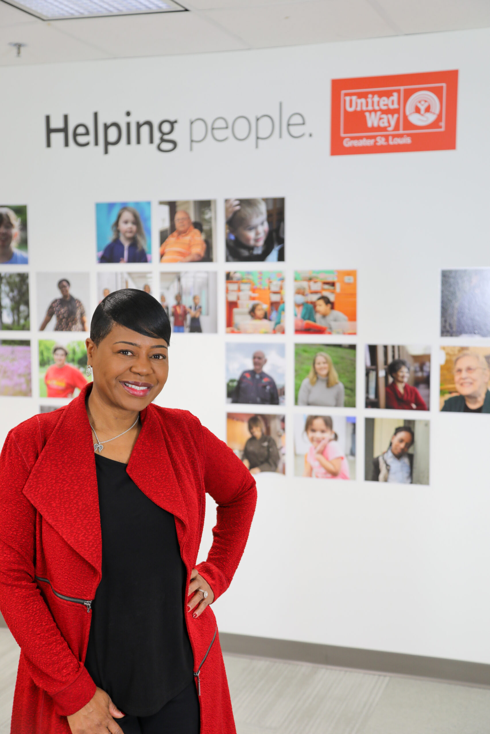 Michelle Tucker, in a red jacket, stands smiling in an office with a wall display behind her. The display features photos of various people and the text "Helping people," along with the United Way Greater St. Louis logo, highlighting volunteer opportunities with this renowned non-profit organization.