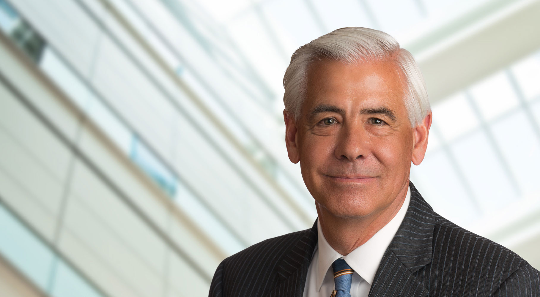 A suited man with white hair smiles in front of a modern building with glass windows and steel beams.
