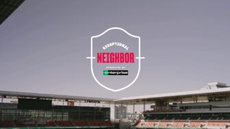 A stadium with empty seats under a clear sky features the words "Exceptional Neighbor presented by Enterprise" prominently in a shield-shaped logo at the center of the image. This scene embodies community spirit and highlights volunteer opportunities, welcoming those ready to make a difference.