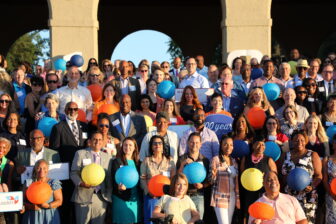 A large, diverse community gathers outdoors, many holding colorful paper lanterns and smiling at an event beneath arches and trees. Some hold signs with writing on them, showcasing a united way of celebrating together.