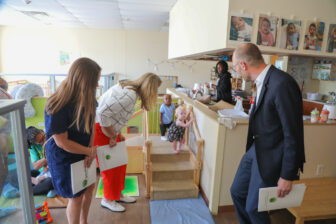 In a lively daycare setting, a small group of adults engages with children, fostering community connections. A toddler climbs stairs under the watchful eyes of two crouching women, while a man in a suit observes nearby. The room is filled with colorful decor and play areas, emphasizing volunteer opportunities.