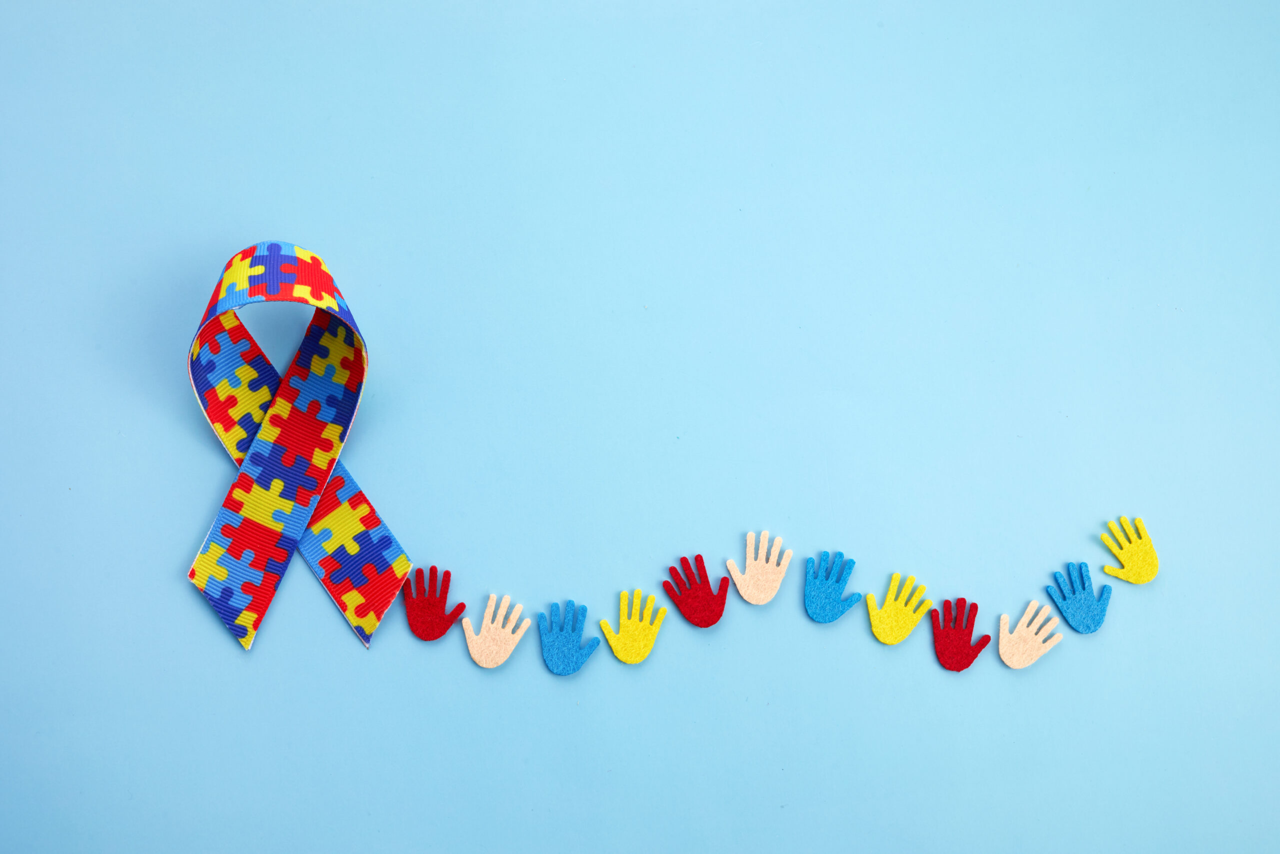 The image shows an Autism Awareness ribbon made up of multi-colored puzzle pieces. There are several paper cut-out hands trailing after the ribbon.